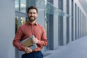 Professional businessman smiling outside urban office holding tablet and notebook. Confident expression, casual attire. Ideal for business, career advancement, confidence themes. photo