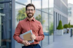 A man wearing glasses and a red shirt is holding a laptop and a piece of paper. He is smiling and standing in front of a building photo