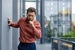 Businessman coughs in outdoor office environment, evoking themes of health, stress, and respiratory concerns. Image signifies focus on wellness and potential illness during workday. photo