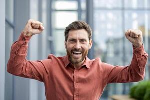 Happy man with raised fists celebrating success in office environment. Joyful expression, relaxed casual attire signify confidence, motivation, achievement, victory, positivity. photo