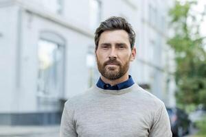 A man with a beard and a blue shirt is standing in front of a building. He is wearing a gray sweater photo