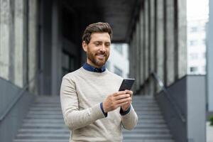 Confident man holding phone, smiling as he stands in front of corporate building. Represents business connectivity, technology, and communication. Ideal image for depicting professional lifestyle photo