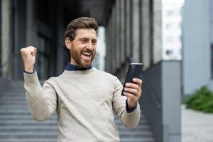 Man celebrating success while looking at smartphone. He is expressing happiness and excitement with fist pump. Urban background adds dynamic energy to his joyful reaction. photo