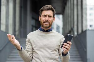 Businessman with questioning look holds phone in urban office environment showing confusion. Adult male thinking outside modern building demonstrates emotion and problem-solving concept. photo