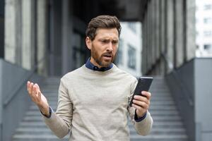 Businessman standing outside, looking frustrated at smartphone. Displaying expressions of confusion and stress. Depicts common challenges of modern work life. photo