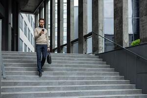 Businessman descends urban stairs while focused on smartphone, demonstrating modern professional life. Dressed in casual business attire, he efficiently multitasks between phone photo