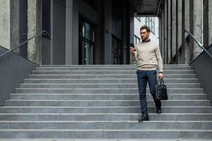 Confident businessman descending stairs holding phone and briefcase. Wearing glasses, sweater, and trousers, he appears focused and professional. Scene captures modern business lifestyle and urban environment. photo