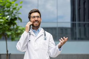 Confident doctor in white coat with stethoscope and glasses talking on phone outside modern building. Professionalism and effective communication emphasized through friendly expression. photo