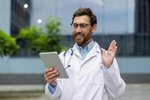 Medical professional in white coat with stethoscope engaging in pleasant call using tablet. Displays easy communication and telemedicine. Portrays modern healthcare and outdoor setting. photo