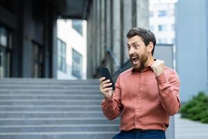 Man with phone in hands received online notification message about winning win. Businessman celebrating successful results achievement from outside office building, holding hand up. photo