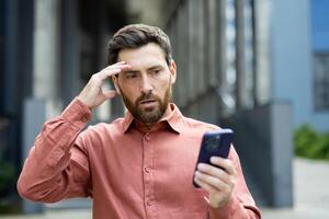 Upset and disappointed man with phone in hands, businessman received online notification message with bad news, walking outside office building, using app. photo