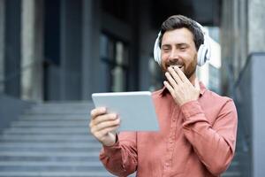 A man wearing headphones and holding a tablet computer is watching a funny and entertaining online stream while walking outside an office building. photo