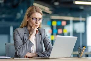 Confident businesswoman using laptop in office. She is deep in thought while analyzing data for work projects. Professional attire and focused expression highlight her commitment to business goals. photo