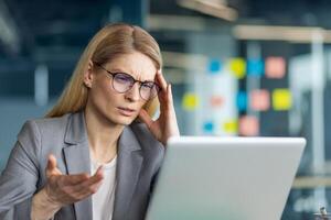 Frustrated businesswoman with glasses using laptop in office. She appears confused and stressed while working on computer. Professional attire suggesting corporate environment photo