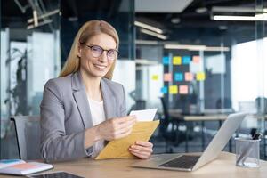 Mature woman businesswoman holding an envelope and smiling while working in a contemporary office. The workspace features a laptop and organizational tools, creating a productive work setting. photo