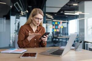 Upset and disappointed woman sitting inside office with phone in hands, businesswoman received online notification message with bad news, app error and phone locked. photo
