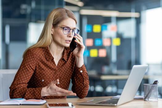Confident businesswoman with engaged in phone call while working at desk in contemporary office. Focused on conversation, utilizing laptop and notepad for efficient multitasking during workday. photo