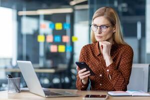 Professional woman in business attire using smartphone and laptop at office desk. Concentrated on planning and decision-making, exploring technology for efficient work. photo