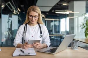 Mature female doctor checks digital tablet at desk in bright office. Equipped with stethoscope, uses technology for efficient healthcare management. Professional setting with laptop and documents. photo