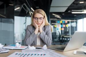 Sad disappointed woman sitting near unfinished financial report. Financier inside office, working with documents and graphs. Using laptop in paperwork. photo