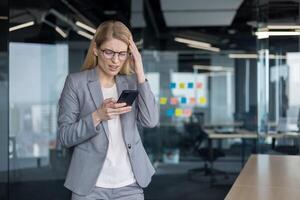 Bad news, negative online notification. Woman holding phone in hands disappointed and sad received message error using app. Businesswoman inside office at workplace nervous irritated. photo