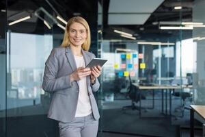 Portrait of a successful mature businesswoman with a tablet computer in her hands. Office worker in a business suit smiling and looking at the camera, standing near the window inside the office. photo