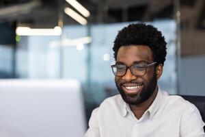 African American businessman smiling while working on computer in modern office. Glowing screen reflects focus, productivity. Professional attire highlights engagement, technology use. photo