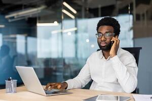 Portrait of serious confident businessman, man thoughtfully looking at camera, working inside office at workplace with laptop, talking on phone. photo