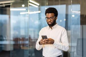 Serious focused man standing inside office with phone in hand, businessman typing text message thoughtfully, employee in business clothes using app at workplace. photo