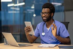 African American male healthcare professional using tablet for virtual consultation at office desk. Displaying telemedicine practices with laptop, documents, digital communication, and connection. photo