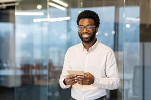 Portrait of a young successful businessman inside the office at his workplace. The man is smiling and looking at the camera, holding a tablet computer in his hands. photo