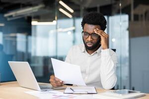 Tired and overworked man with paperwork inside office. Office worker dissatisfied with financial report. Businessman holding document, tax statistical form in hands. photo