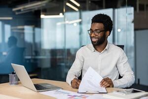 Young successful financier doing paperwork inside office. Man smiling happily, working with laptop and documents, graphs and charts. Businessman in shirt preparing report. photo