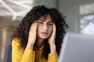 Headache, woman is overloaded and overtired at workplace inside office working with laptop. Office worker holds head in hands, looking at computer screen. photo