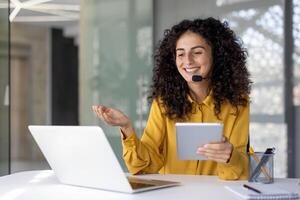 Support service worker, businesswoman with headset and tablet in hands communicates and consults clients remotely, office worker smiles satisfied uses laptop inside office. photo
