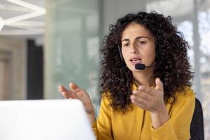 Serious and focused woman with headset talking remotely, close-up of helpline worker working remotely, using laptop for call, at workplace inside office. photo