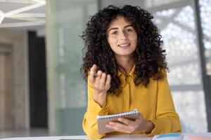 Woman with notebook in hands watching online course, webcam view. Office worker smiling and looking into camera, asking questions and communicating with colleagues remotely. photo