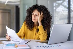 Hispanic businesswoman sits at desk with laptop, reviewing documents with concentrated expression. Image conveys stress, analysis, and multitasking in workplace setting. photo