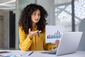 Hispanic businesswoman engaging in call presentation, explaining data analysis while holding paper chart. Confident expression, professional setting, demonstrates expertise in business photo