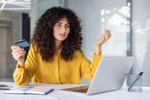 Woman sitting at desk holding credit card while looking at laptop with frustrated expression. Indicates online payment issue or possible fraud, confusion over transaction or account error. photo