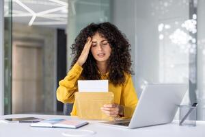 Stressed woman receiving bad news from envelope in office setting. Expression of worry while seated at desk with laptop, phone, and various office essentials. Modern professional environment. photo
