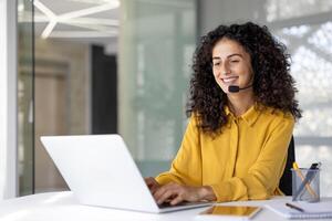 Joyful customer support and consulting service worker with headset smiling, using laptop for call. Woman inside office, remotely helping customers, supporting. photo