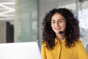 Joyful customer support and consulting service worker with headset smiling, using laptop for call. Woman inside office, remotely helping customers, supporting. photo