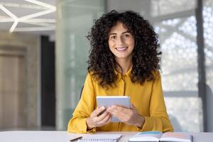 Happy and successful woman sitting at desk with tablet computer. Businesswoman at workplace inside office smiling and looking at camera, using financial app. photo