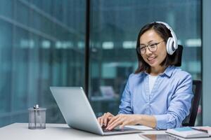 Young Asian programmer with headphones working inside office with laptop, businesswoman at workplace typing on keyboard, listening to music, coding new software. photo