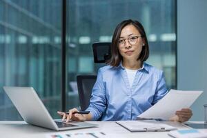 Asian businesswoman working with documents and a laptop in a modern office setting. She holds a professional demeanor, clearly showcasing her focus and dedication to task. photo