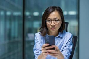 Serious focused woman using app on phone, Asian thinking businesswoman close up typing message on smartphone, working inside office near window. photo
