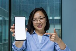 Asian businesswoman holding a smartphone with a blank screen, pointing towards it and smiling. Perfect for representing technology, business presentations, or mockups for app design and development. photo
