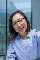 Asian woman smiling and looking at the phone camera, woman inside the office at the workplace working and talking to colleagues and friends using an application on a smartphone. photo