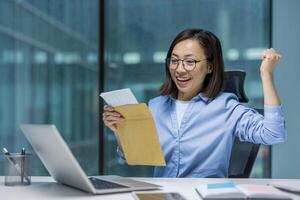Joyful and successful Asian woman at workplace received mail notification letter envelope with good winning news, businesswoman sitting with laptop, holding hand up reading message. photo